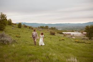 Two people in wedding attire walk down a grassy hillside overlooking a scenic mountain valley on an overcast day.