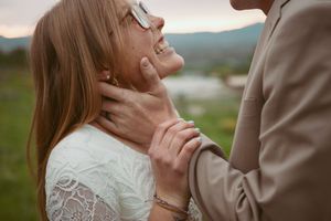 A close-up romantic moment showing hands gently holding a face against a blurred mountain landscape background.
