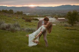 A couple in wedding attire share a romantic dip pose on a hillside at sunset with mountains and a lake in the distance.