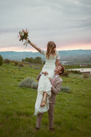 Romantic couple in formal wear dance and embrace on a grassy hillside overlooking a scenic mountain landscape at sunset.