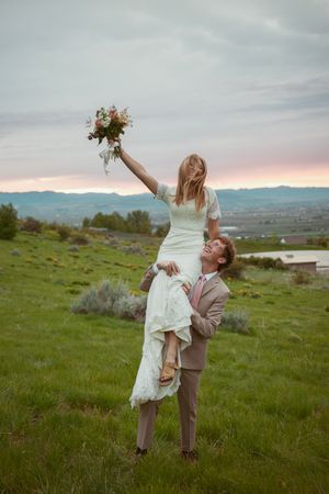 Newlyweds playfully pose in a grassy field with mountain views at sunset.