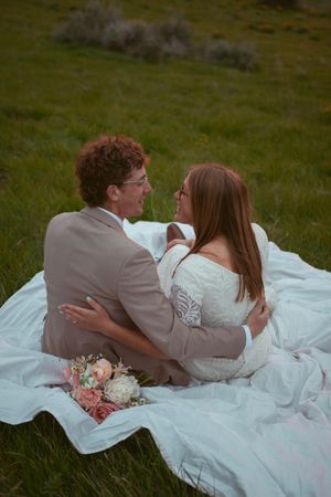 A couple in wedding attire sits together on a white blanket in a grassy meadow with a pink floral bouquet nearby.