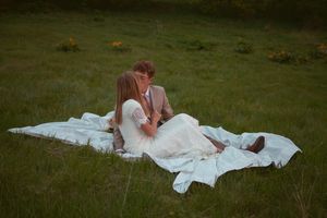 A couple in wedding attire lies on a white blanket in a grassy field.