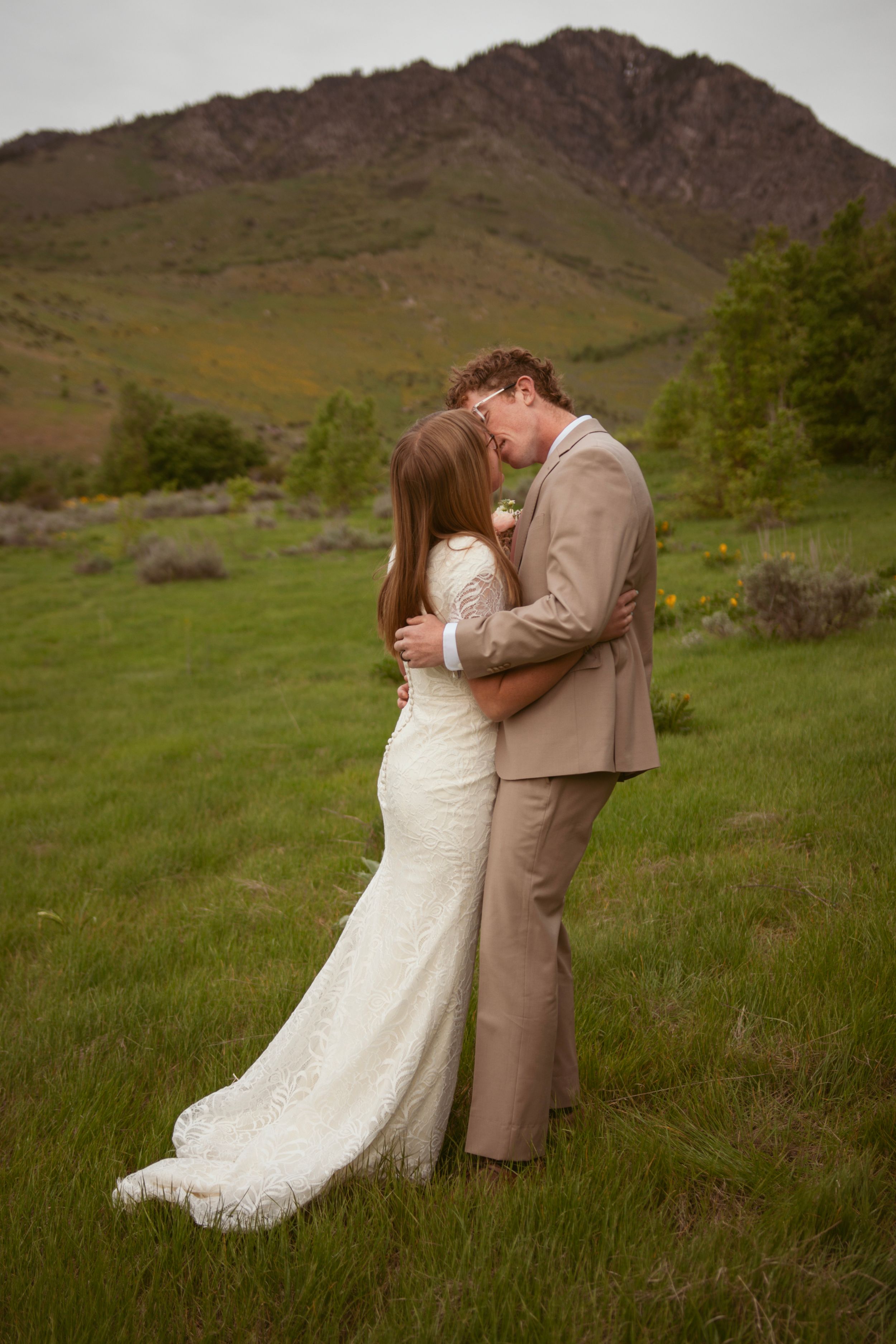 A romantic couple dips dramatically on a hillside meadow with mountains in the background during a wedding photo shoot.