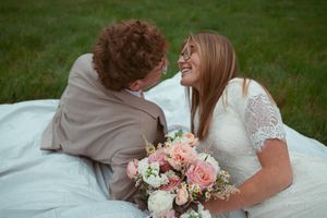 Tender moment between newlyweds with delicate pink and white bouquet on grassy hill.