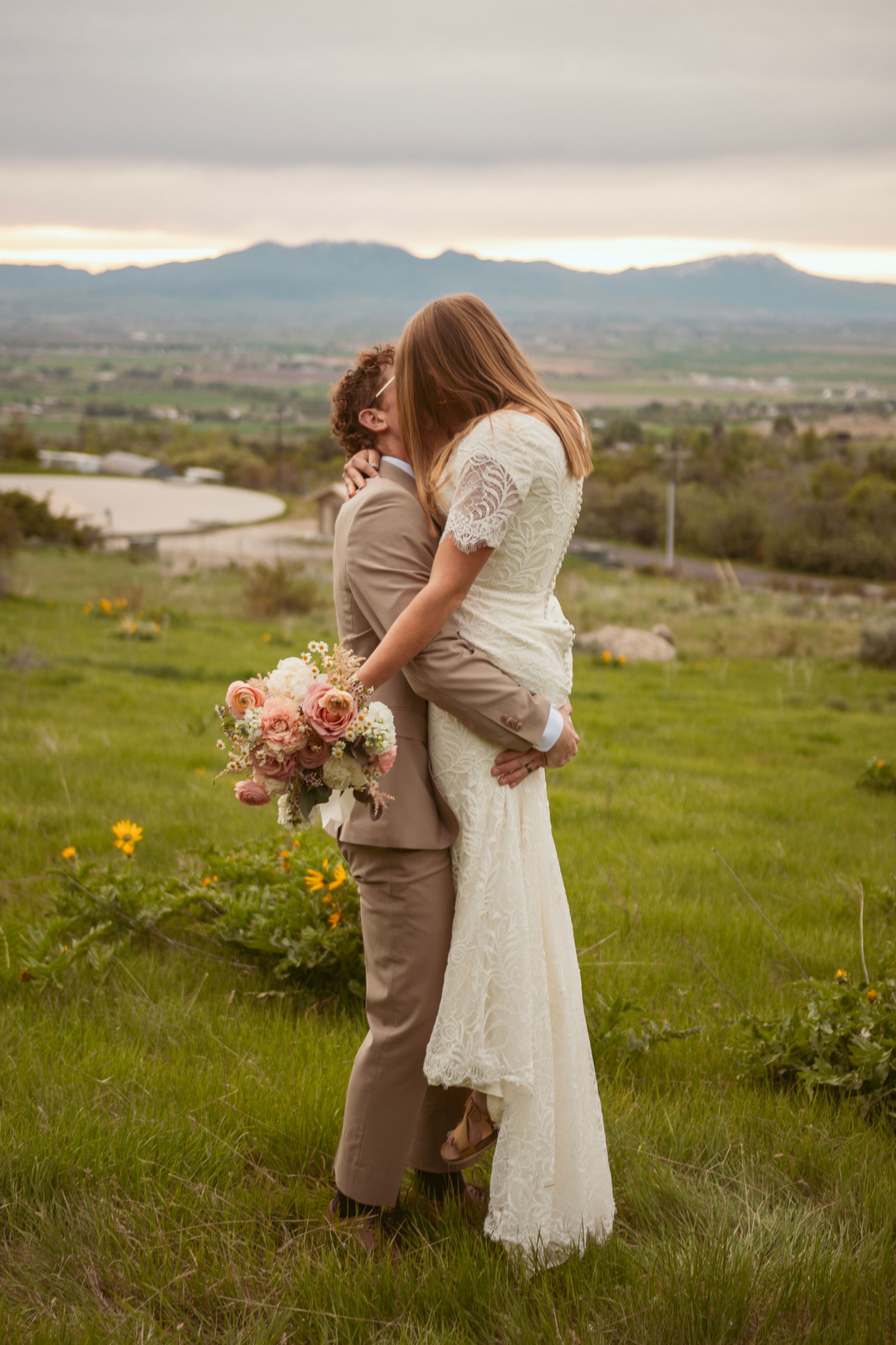 Couple in wedding attire shares an intimate moment in a wildflower-dotted meadow with mountain views.