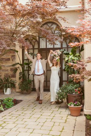 A joyful couple celebrates with raised arms under arched doorways and Japanese maple trees.
