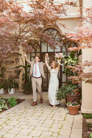 Joyful newlyweds with raised arms celebrate under autumn foliage at elegant stone courtyard entrance.
