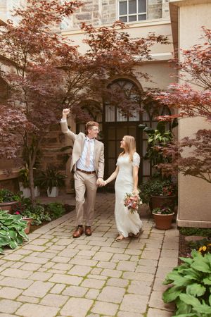 A couple in wedding attire playfully poses and dances together on a stone patio with Japanese maple trees overhead.