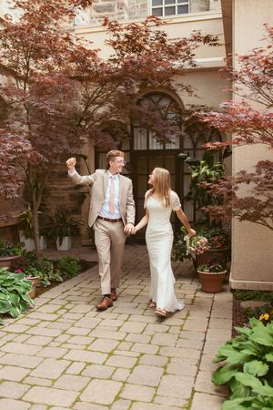 A couple walks hand in hand on a stone path surrounded by Japanese maple trees and garden planters.