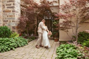A couple embraces on a stone path surrounded by red maple trees and green hostas near an arched doorway.