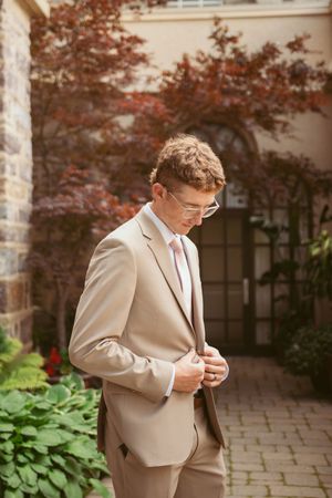 A groom in beige suit adjusts his tie while standing on a stone patio surrounded by greenery and brick walls.