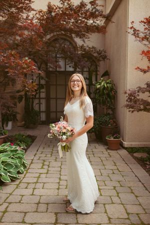 Bride in white lace gown holds pink bouquet while standing in garden courtyard with maple trees.
