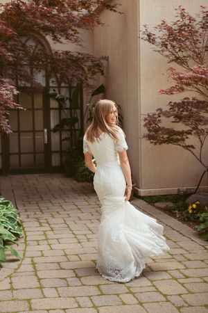 A bride in a fitted lace gown poses on a stone walkway framed by Japanese maple trees.