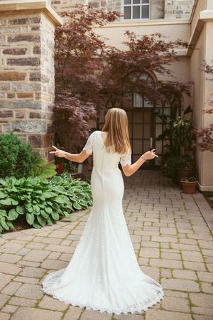 A bride in an elegant lace mermaid wedding dress stands on a stone pathway beside blooming hostas and a Japanese maple tree.