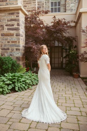 A white lace wedding dress with long train shown from multiple angles on a stone patio with lush garden backdrop.