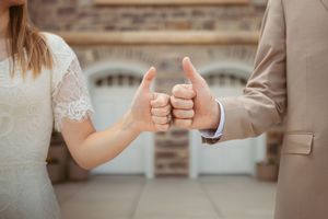 Two people hold hands and give a thumbs up gesture while wearing wedding attire.
