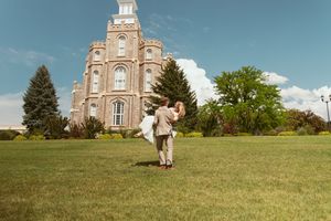 A well-dressed couple stands on a green lawn in front of a grand historic temple with ornate architecture and spires.