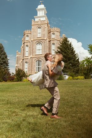 A couple performs a dip dance move on the lawn with a grand stone building featuring a tower in the background.