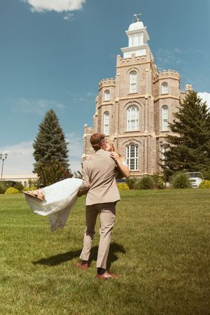 A couple performs an energetic dance lift on a green lawn with a historic stone church as the backdrop.