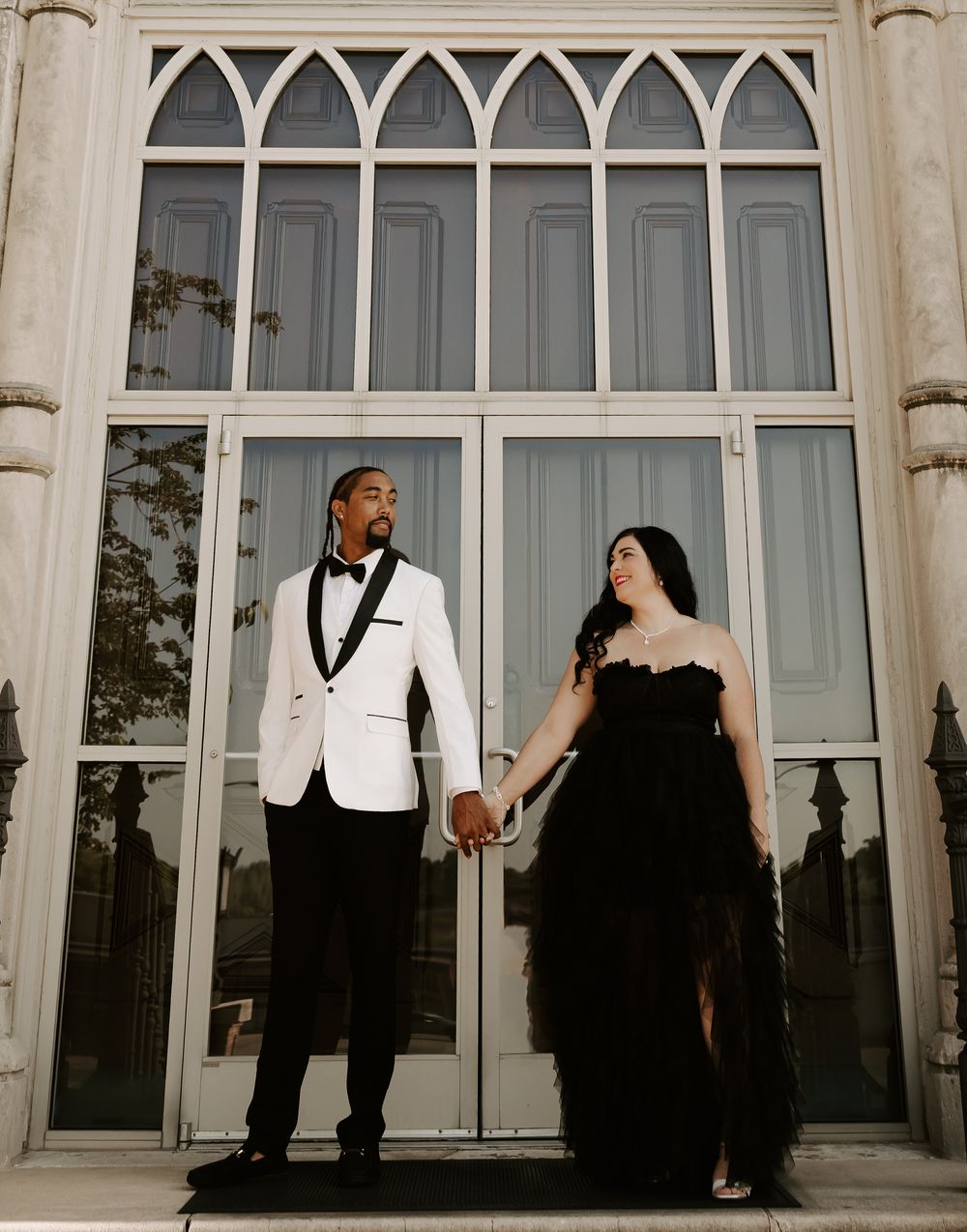 A couple in formal black and white attire poses against a grand building with arched windows.