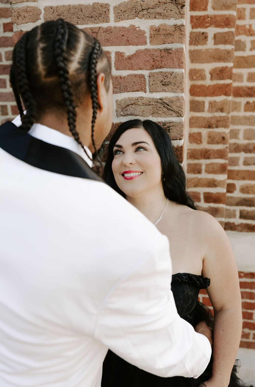 A romantic couple poses against a brick wall, with one wearing a white suit jacket and the other in dark attire.