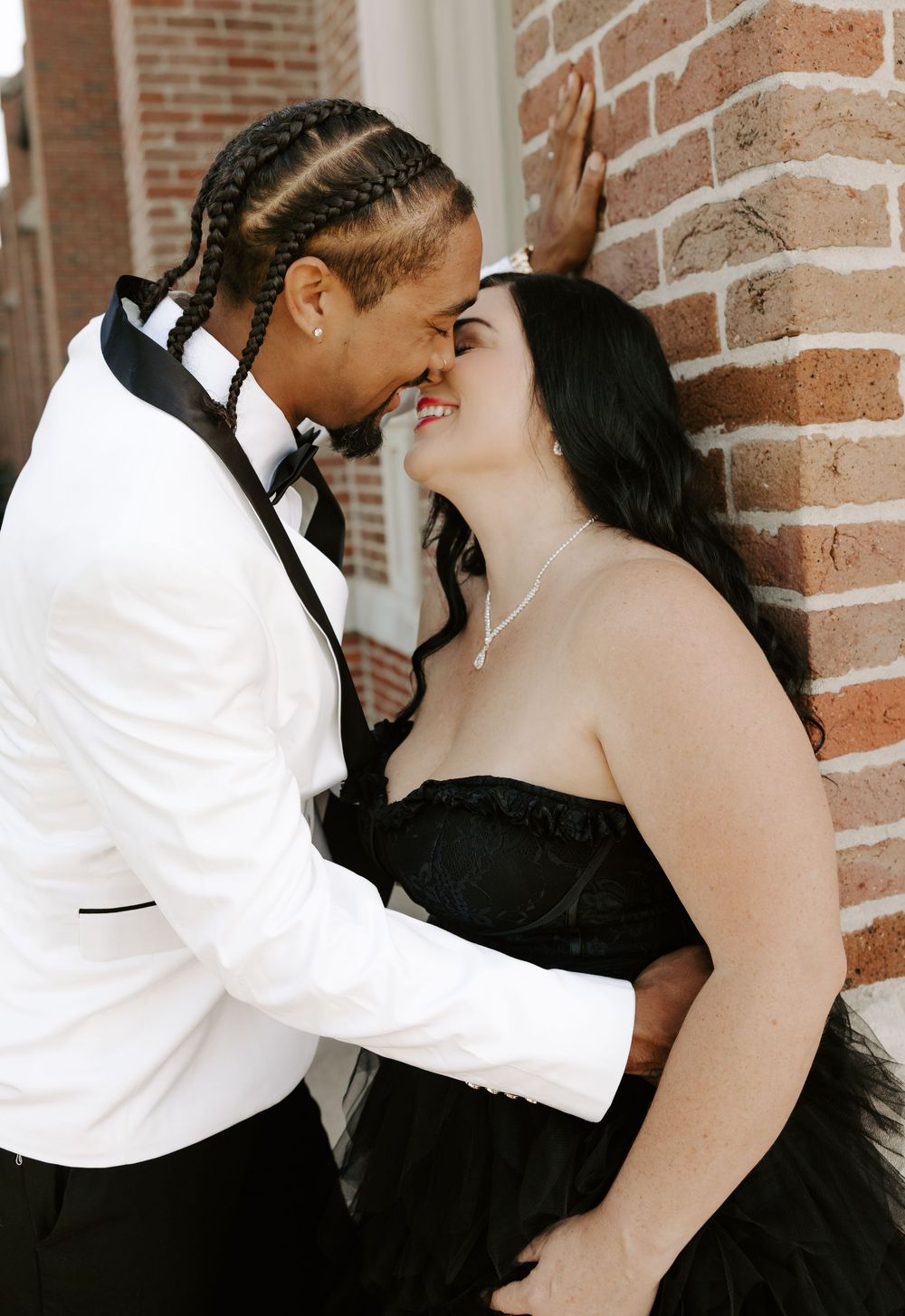 A tender moment captured between a couple in formal wear against a rustic brick backdrop.