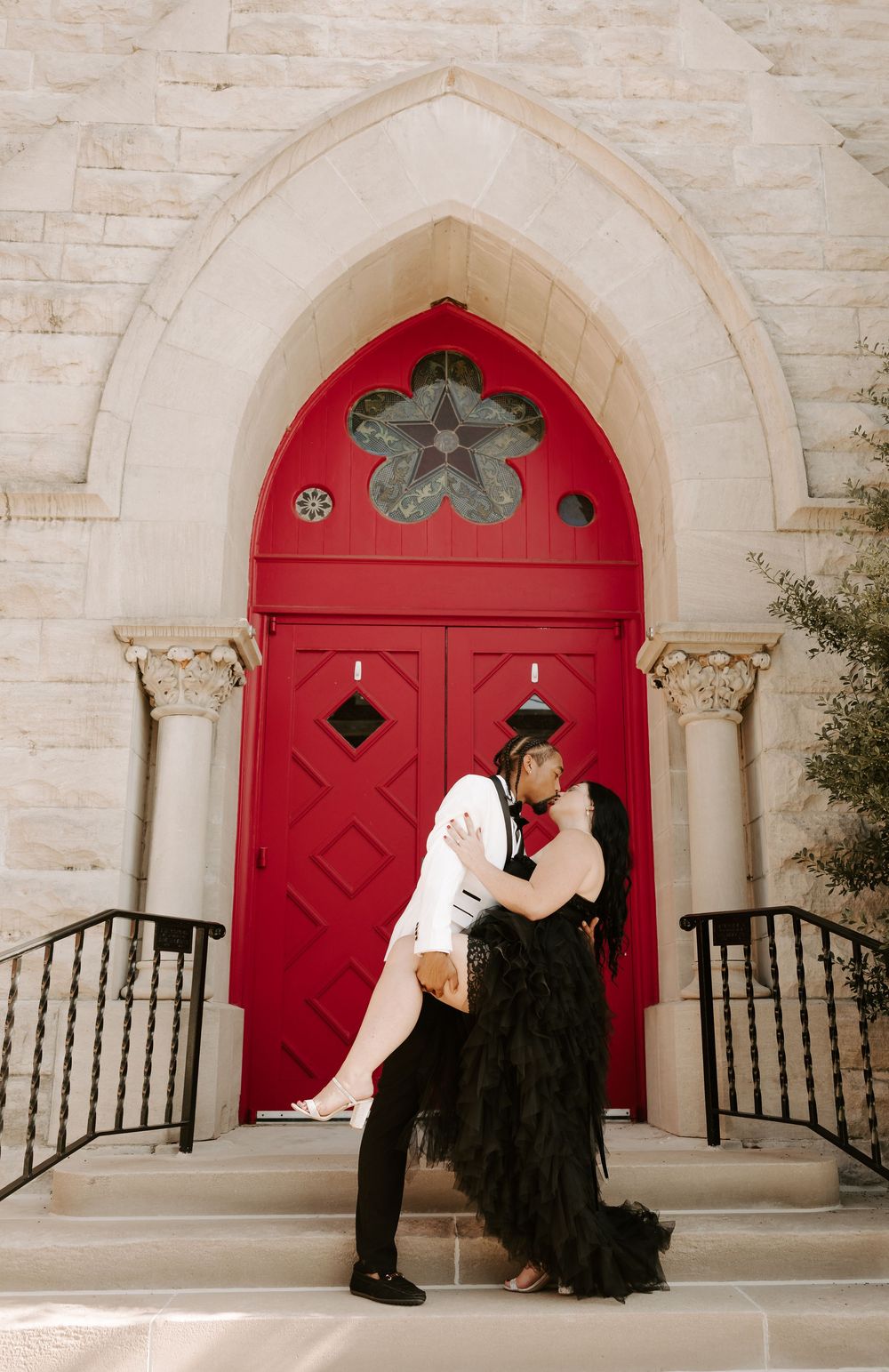 A couple poses dramatically in front of a red church door with gothic architecture.