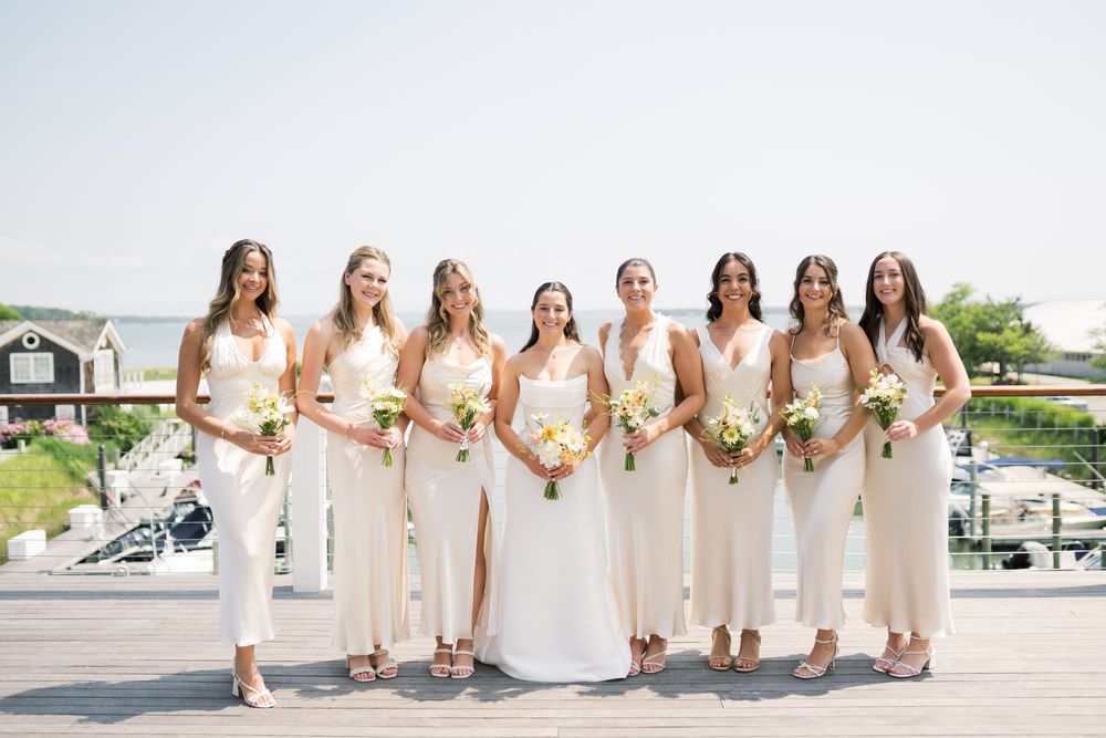 Bridal party in elegant cream dresses poses together on an oceanfront deck.