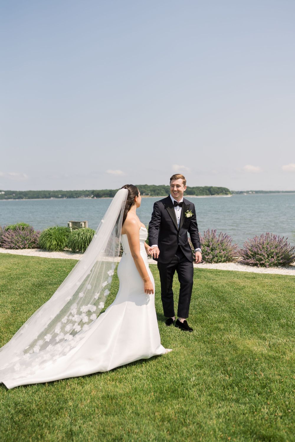 A couple in wedding attire poses on a grassy lawn with a waterfront view and purple flowers in the background.
