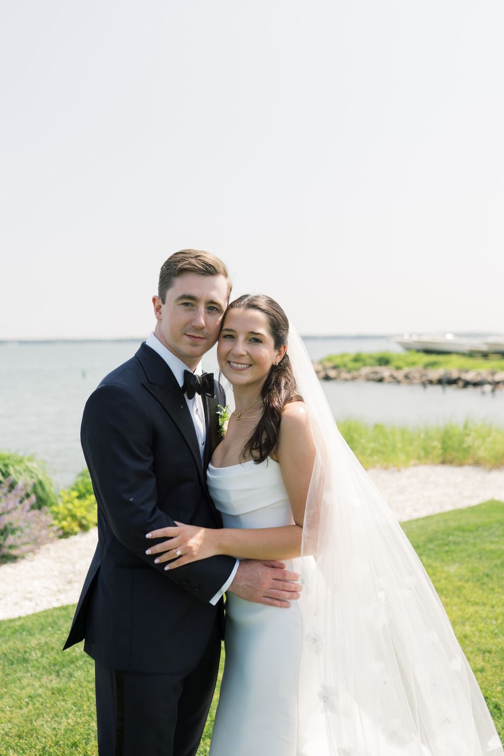 Newlyweds embrace on a scenic waterfront lawn during their wedding photoshoot.
