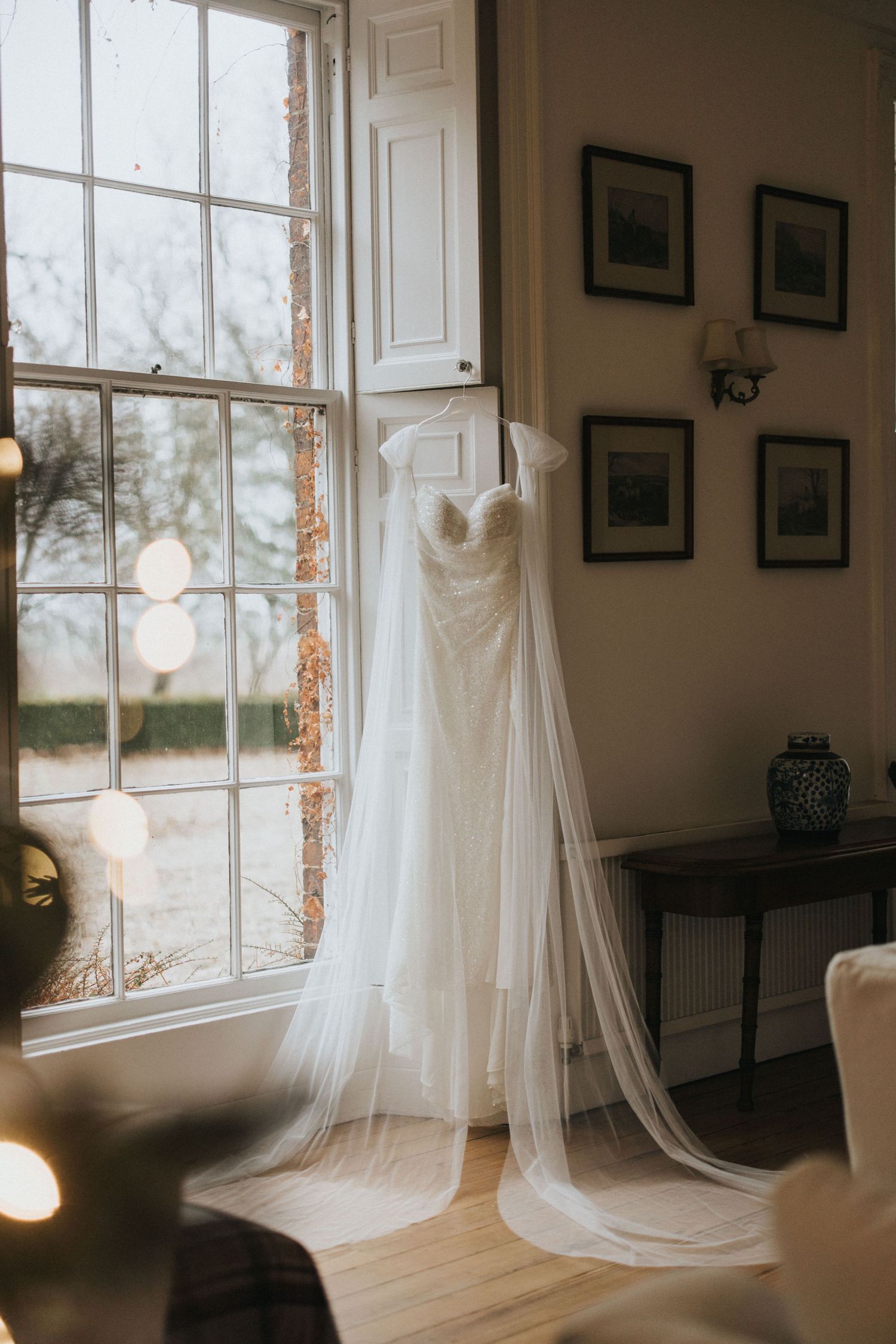 White wedding dresses hanging near sunlit windows in a vintage-style bridal suite interior with framed artwork.