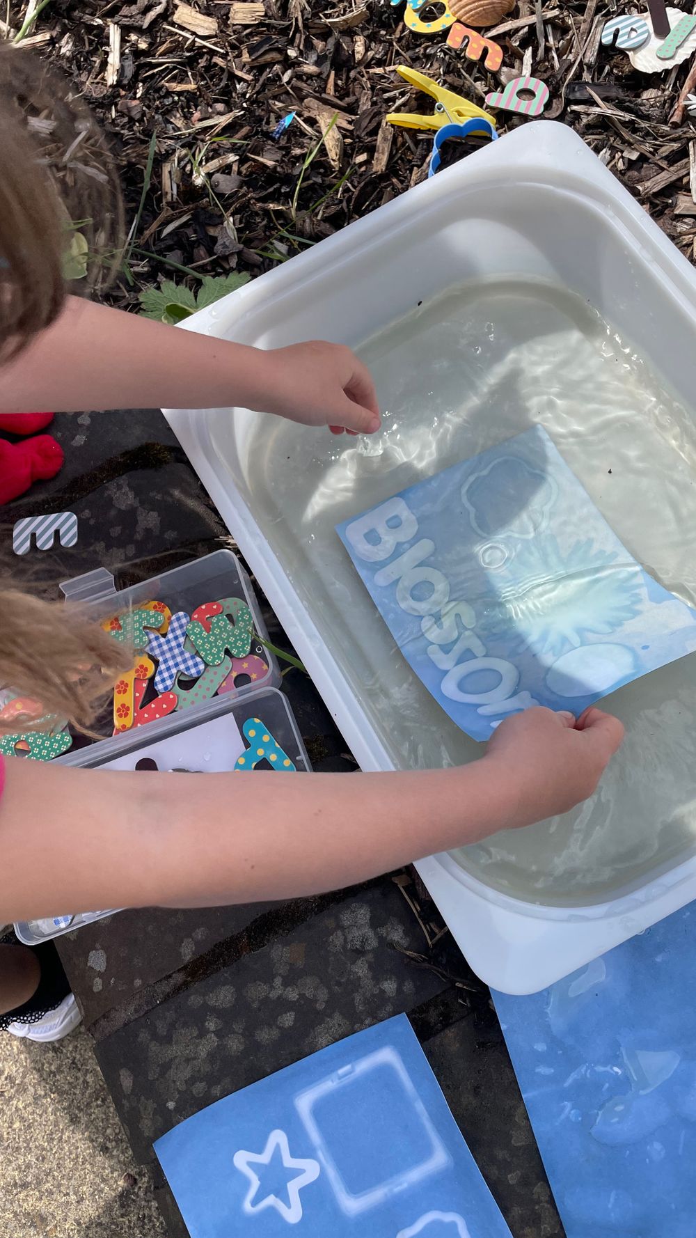 Small hands dipping a blue toy or block into a white container filled with water for sensory play or cleaning.