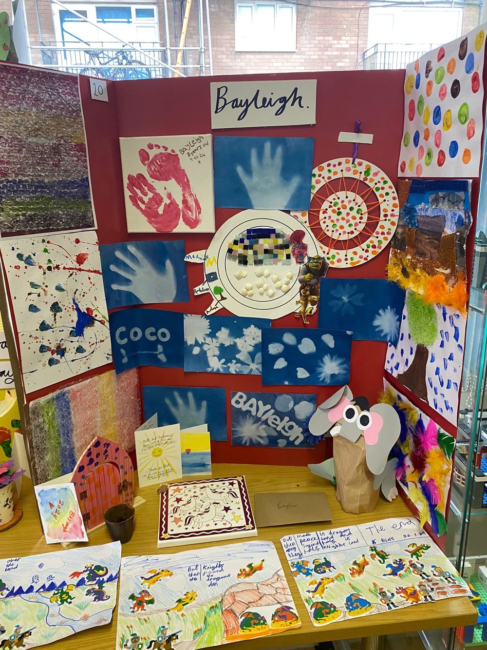 Colorful children's art display board with handprints, drawings and a stuffed bunny toy in an elementary school setting.