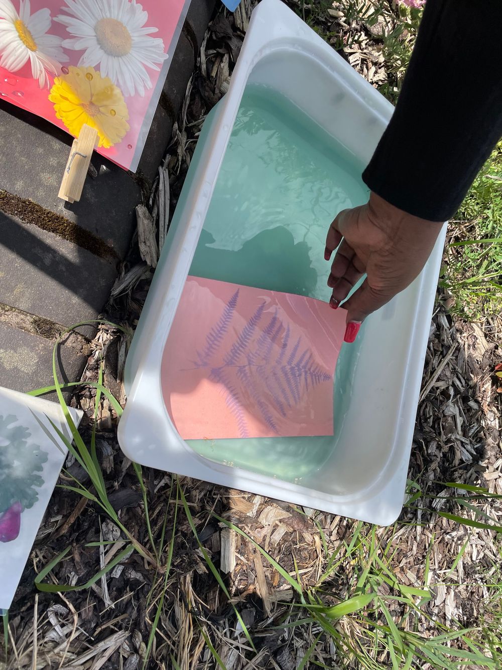 A hand dips paper into a rectangular container filled with turquoise liquid during a crafting activity.