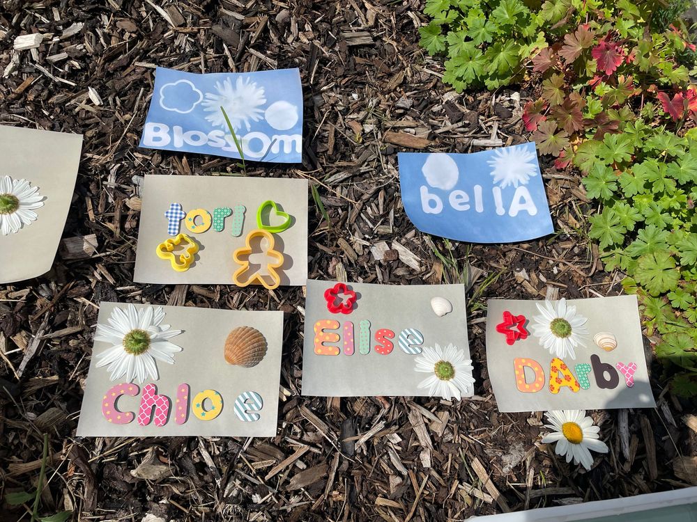 Colorful personalized name plates and decorative tiles arranged on dark mulch in a garden bed with green plants.