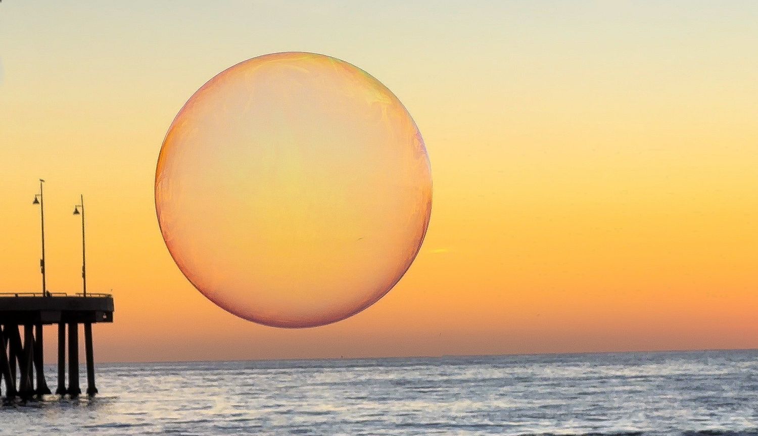 Large orange bubble over ocean pier creates stunning circular silhouette against pastel sky.