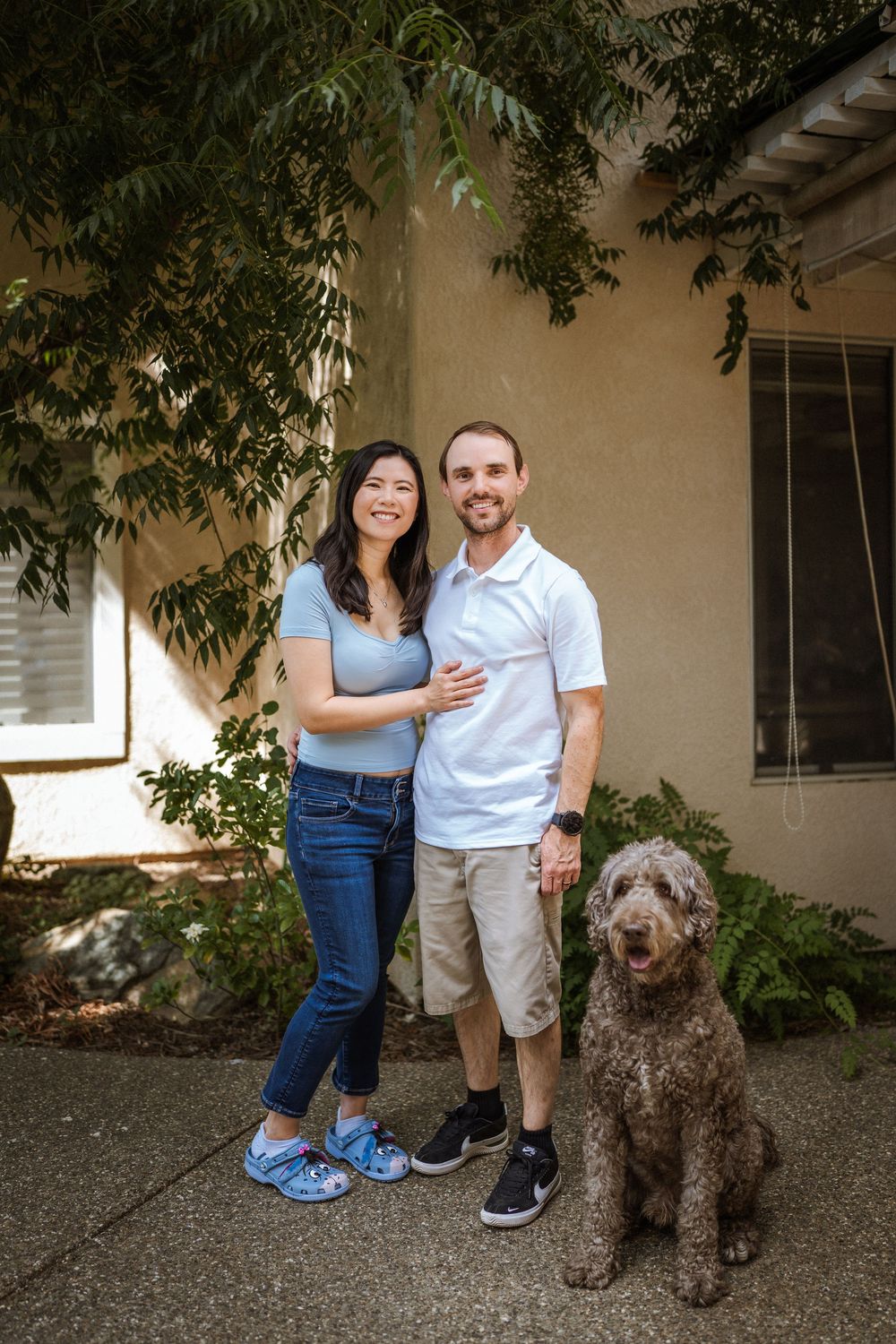 A couple poses with their large brown dog in front of a house with climbing vines.