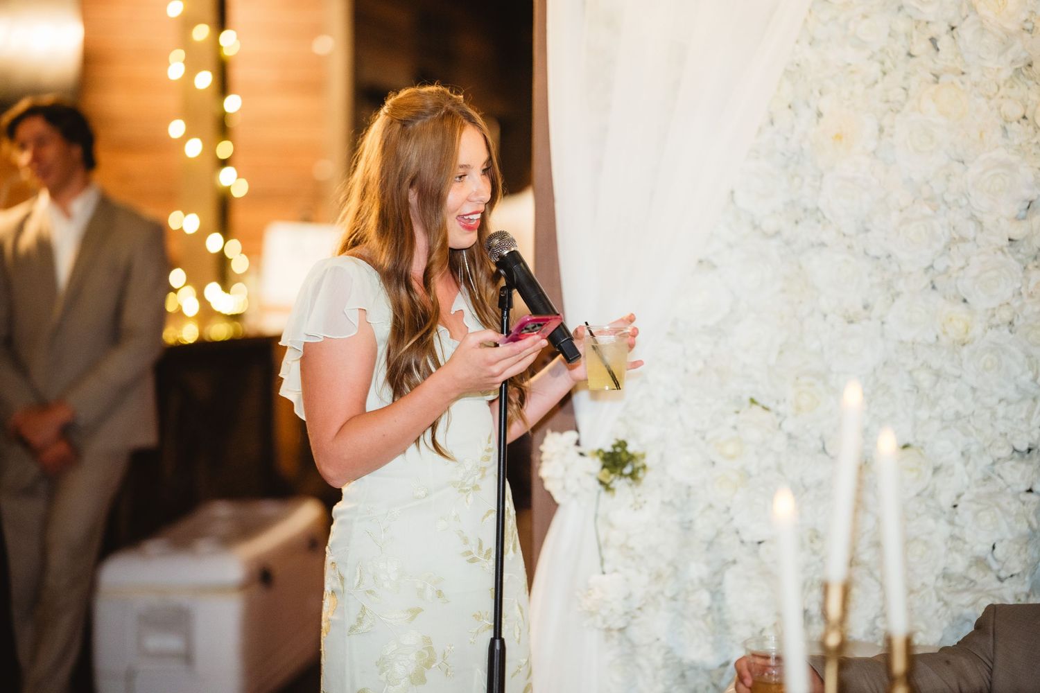 Bride in white dress delivers wedding speech at candlelit reception with bokeh lights in background.