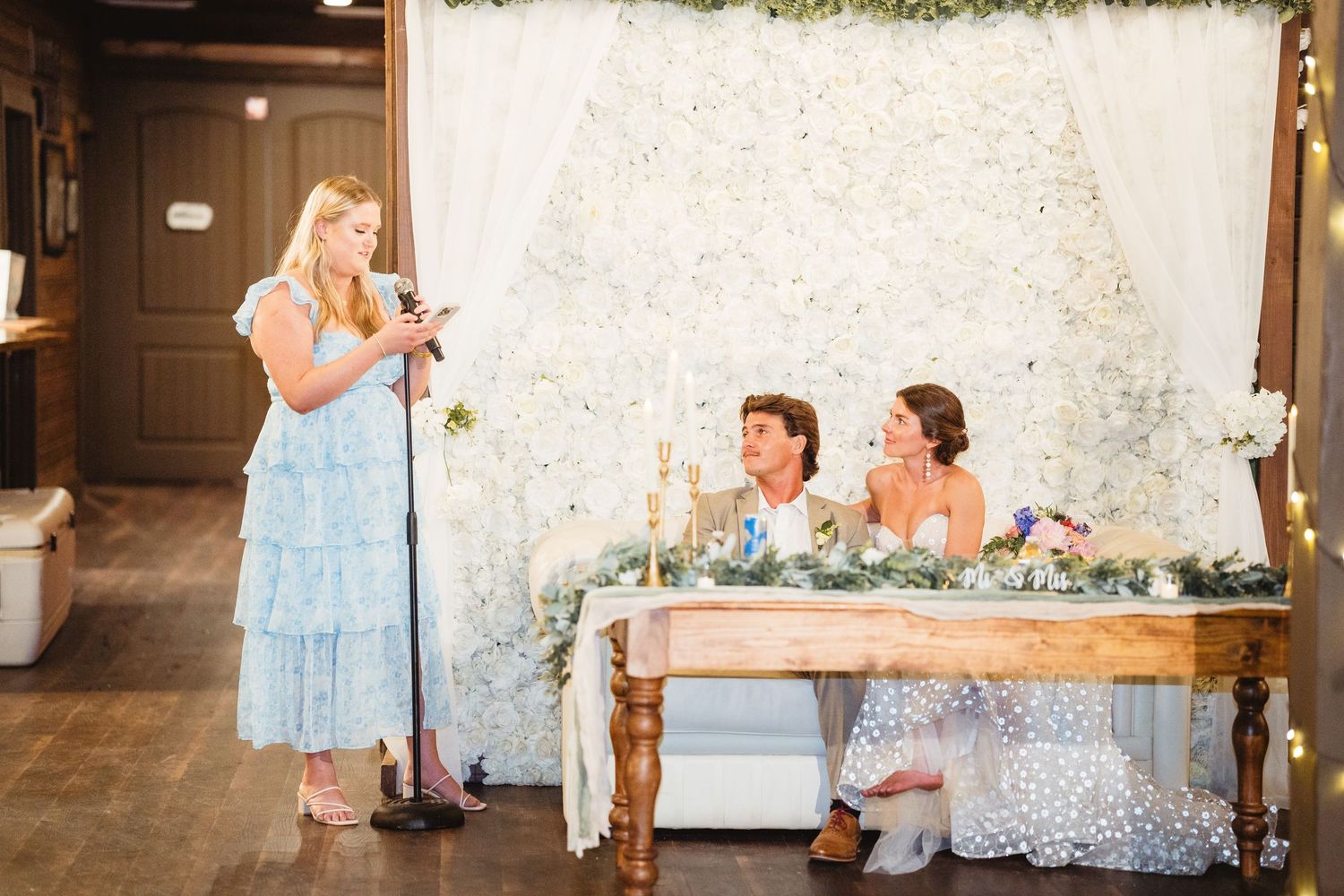 Speech during wedding reception with head table setup and white floral backdrop.