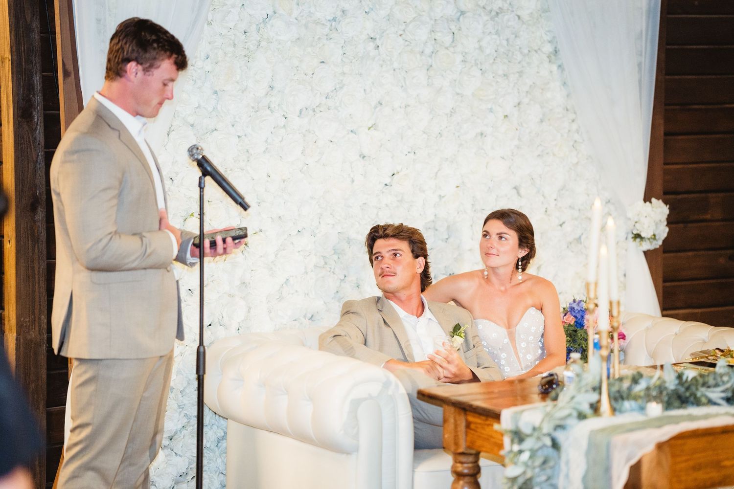 Wedding reception speech with attendees gathered in front of a white textured backdrop.