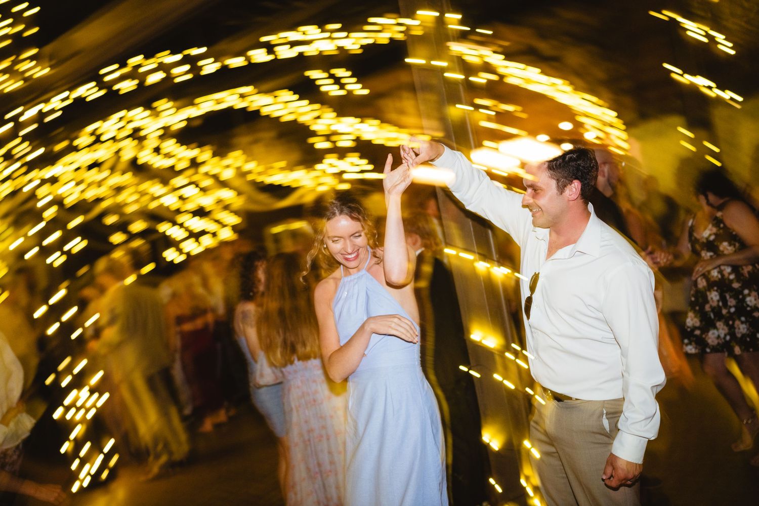 Dancing guests create motion blur streaks under warm yellow venue lighting at a lively wedding reception party.