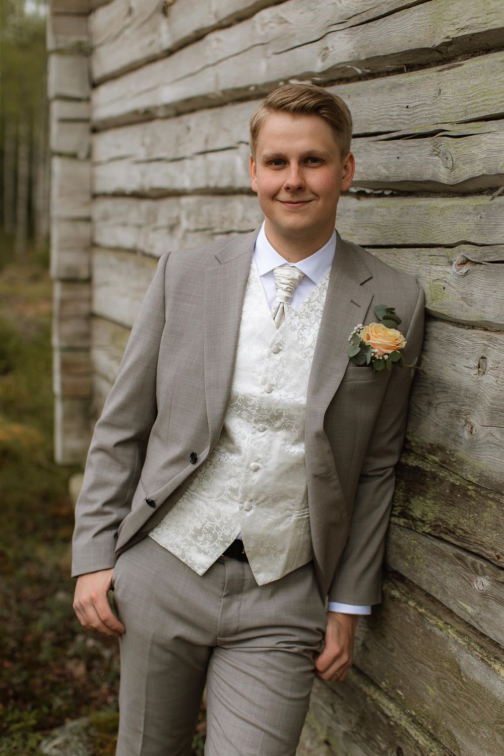En man i grå kostym, klädd för bröllops som står och lutar sig emot en husvägg medan han tittar in i kameran.