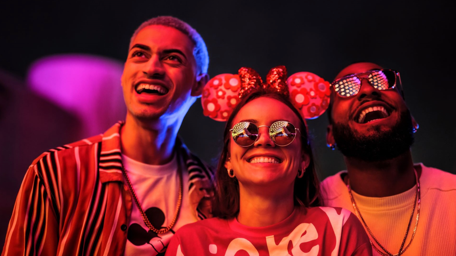 Three park visitors wearing Minnie Mouse ears enjoying a nighttime festival with colorful lighting.