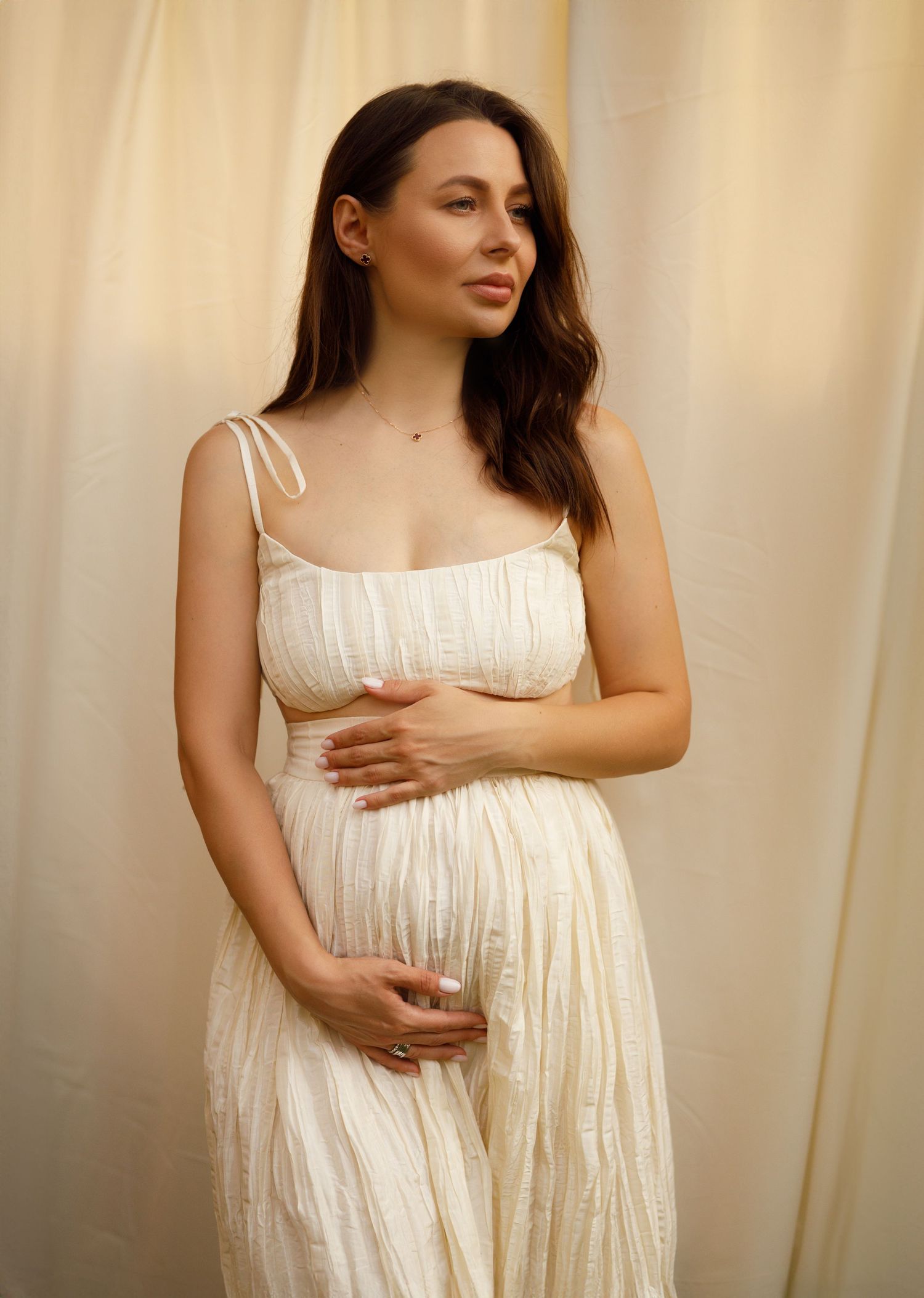 A person wearing a flowing white summer dress poses gracefully against a light-colored backdrop in warm lighting.