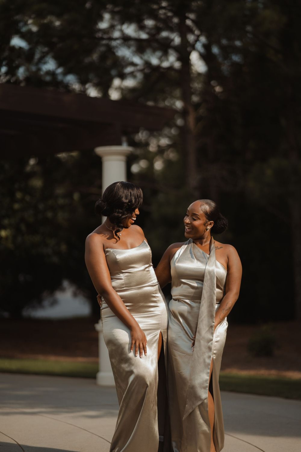 Two people in elegant cream-colored satin dresses share a candid moment outdoors.