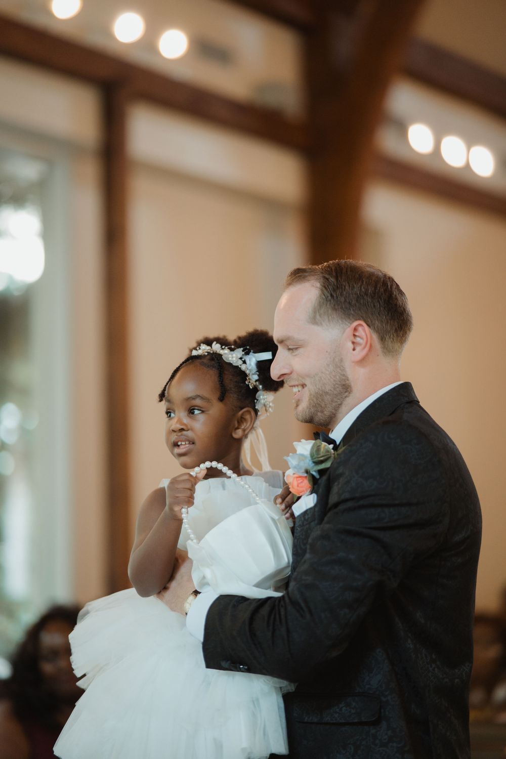 A tender moment shared between a person in a black suit and a young flower girl in a white dress at a wedding celebration.