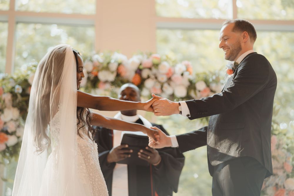A couple holds hands during their wedding ceremony in front of floral decorations and an officiant.
