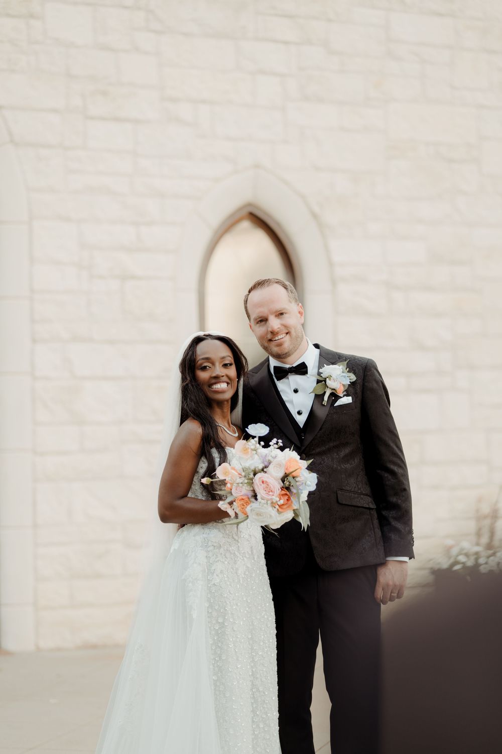 A bride in a white dress and groom in black tuxedo share a joyful moment together against a light stone wall.