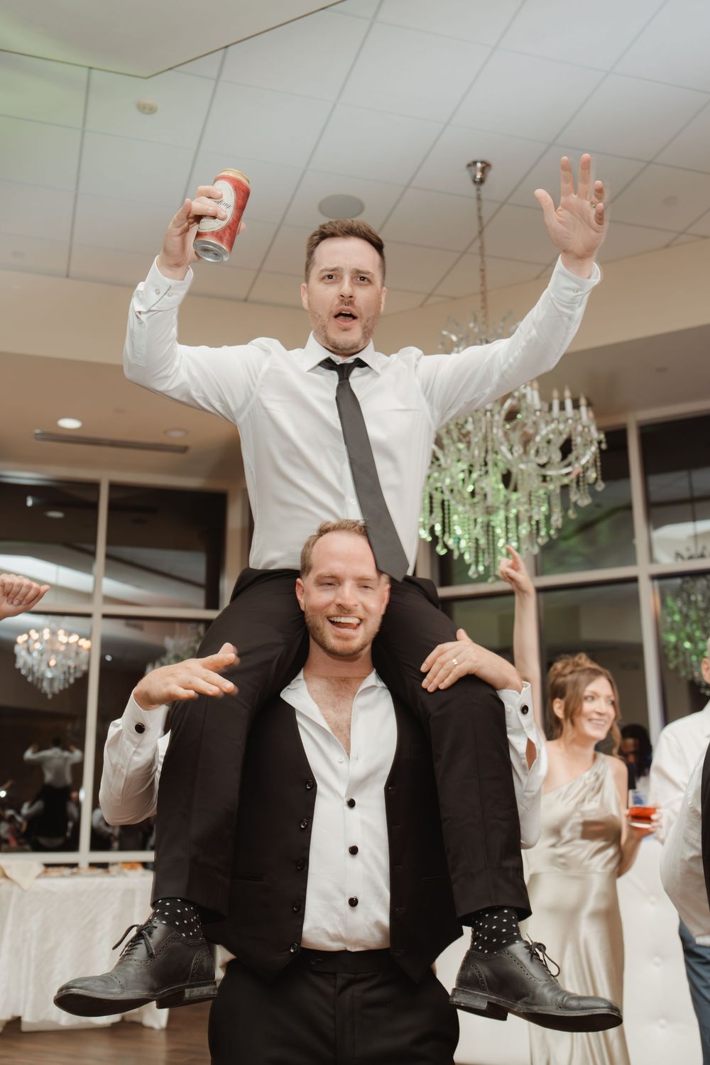 Two wedding guests celebrate on the dance floor with one person riding on another's shoulders while holding a drink.